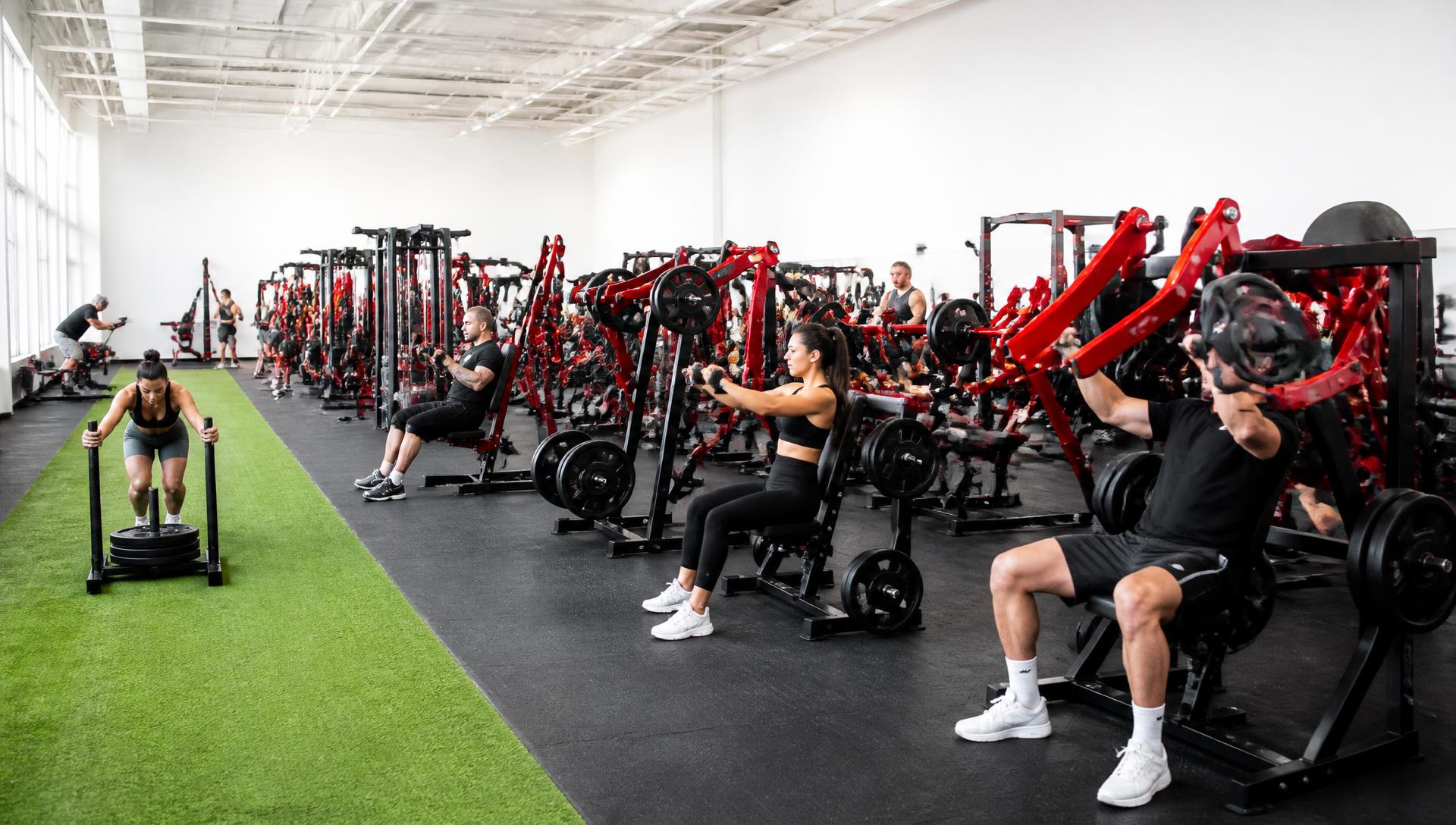Wide view of the Redwood Fitness training floor: rows of red-and-black plate-loaded machines, a green turf lane and members training in daylight